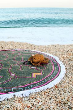 a hat is sitting on top of a round rug at the beach with waves in the background