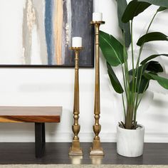 two brass candlesticks next to a potted plant in front of a painting