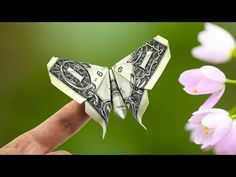 a dollar bill origami butterfly sitting on someone's finger with pink flowers in the background