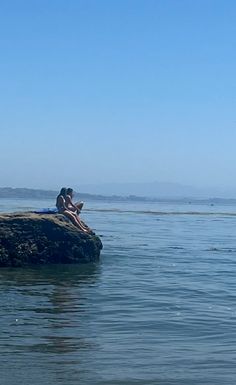 two people are sitting on a rock in the middle of the water looking out to sea