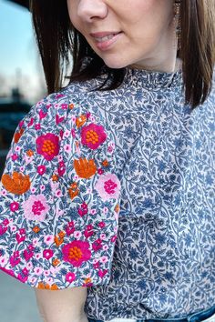 Tight cropped photo of a brunette woman looking off to the side smiling. She is wearing a blue floral woven blouse with a pink and orange embroidered flowers on the puff sleeves. The top also has a mock neck with a small ruffle. Blue Floral Fabric, Front Tuck, The Girlfriends, Twill Shorts