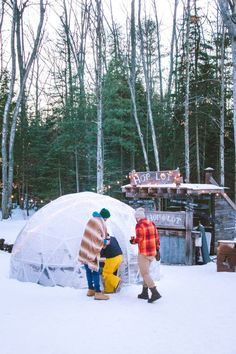 two people standing in the snow next to an igloose with trees behind them