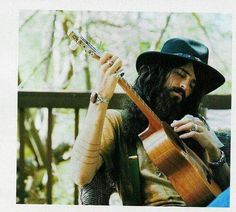 a man with long hair holding a guitar and wearing a black hat while sitting on a bench