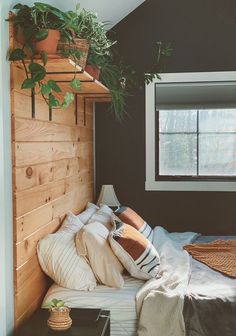 a bed with lots of pillows and plants on the headboard next to a window