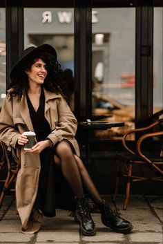 a woman sitting on the ground in front of a building with her legs crossed and holding a coffee cup