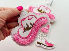 a hand is holding two pink and white patches with gold designs on them, one has a cowgirl's boot in the center