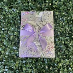 a card with a purple bow on top of some green leaves and bushes in the background