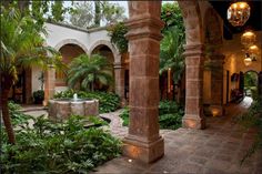 an outdoor courtyard with plants and lights on the ceiling, surrounded by stone pillars that lead to a fountain