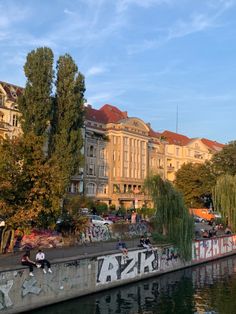 people sitting on the edge of a river in front of some old buildings and trees