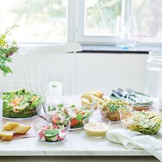 a table topped with lots of salads and bowls filled with food next to a window