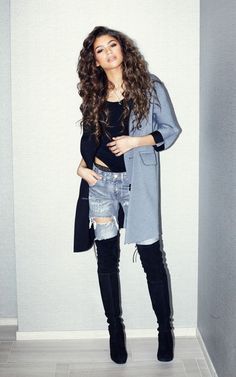 a woman standing in front of a white wall wearing black boots and a gray coat