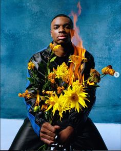 a man sitting on the ground with flowers in front of him and fire coming out of his face