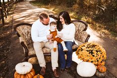 a man and woman sitting on a bench with a baby in their lap, surrounded by pumpkins and gourds