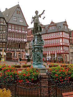 a statue is in the middle of a flower garden with many buildings behind it and a fence around it