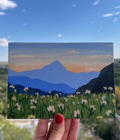 a hand holding up a piece of art with mountains in the background and flowers growing on it