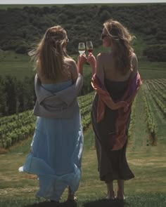 two women standing in the grass holding wine glasses and looking at each other with hills in the background