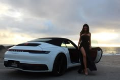 a woman standing next to a white sports car on the beach near the ocean at sunset