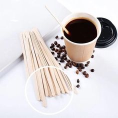 a cup of coffee next to wooden sticks and beans on a white surface with a black lid