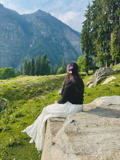 a woman sitting on top of a large rock next to a lush green forest covered hillside