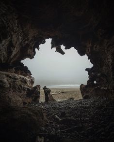 two people are standing in the cave looking out at the beach and ocean on a foggy day