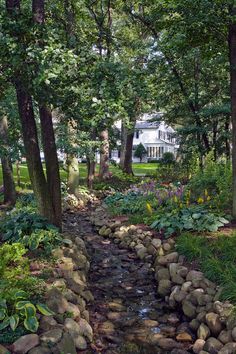 a stream running through a lush green forest filled with lots of trees and rocks in front of a white house