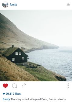 an old black house sitting on the side of a hill next to water and hills