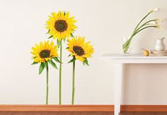 three yellow sunflowers sitting on top of a table next to a white wall