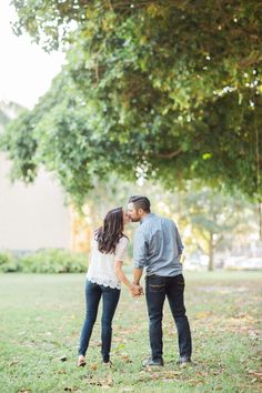 an engaged couple standing in the grass under a tree at their engagement photo shoot by person