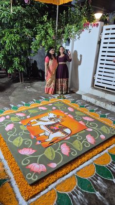 two women are standing under an umbrella in front of a painting on the ground with flowers