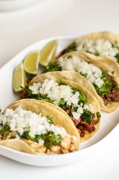 three tacos with meat, cheese and cilantro on a white plate next to lime wedges