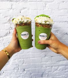 two people holding up green smoothies in front of a white brick wall with the words bowl d on it