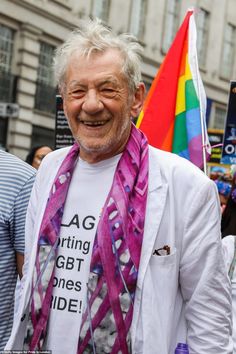 an older man with white hair and wearing a purple scarf smiles at the camera while holding a rainbow flag