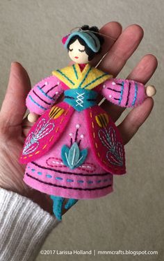 a hand holding a small doll in it's right hand and wearing a pink dress