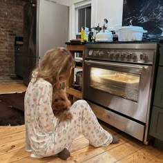 a woman sitting on the floor in front of an oven holding a teddy bear and looking at it