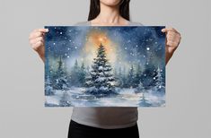 a woman holding up a painting of a snowy christmas tree with the sun in the background