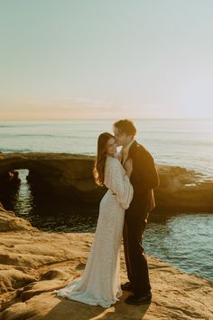 a bride and groom kissing on the rocks by the water at sunset in front of an ocean