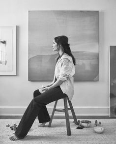 a woman sitting on top of a wooden chair in front of a wall with paintings