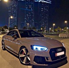 a white car parked in front of a tall building at night with city lights behind it