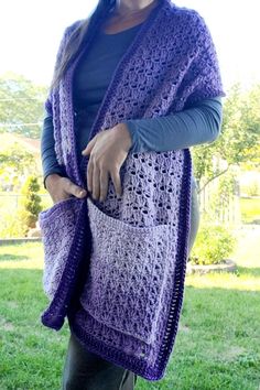 a woman standing in the grass wearing a purple crocheted shawl with pockets