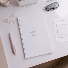 a white desk topped with a notebook and pen next to other office supplies on top of it