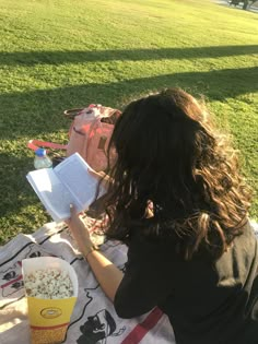 a woman reading a book while sitting at a picnic table with popcorn and drink in front of her