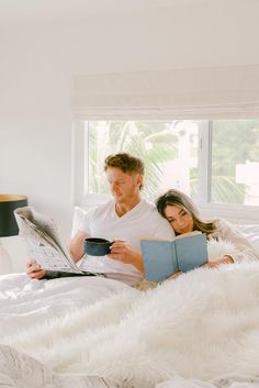 a man and woman laying in bed while reading a book with a cup of coffee