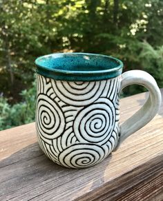 a white and blue coffee cup sitting on top of a wooden table next to trees