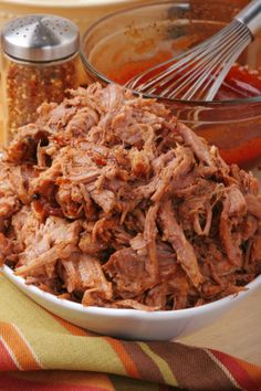 a white bowl filled with shredded meat next to a spoon and seasoning bottle on top of a table