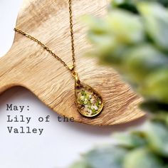 a close up of a necklace on a wooden board with plants in the background and text that reads may lily of the valley