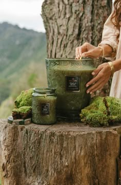 a woman is holding a green container on top of a tree stump with moss growing around it