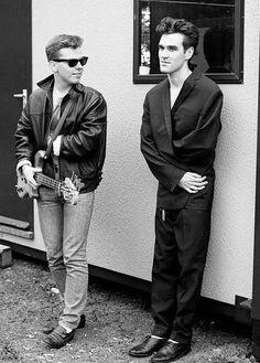 two men standing next to each other near a wall with a guitar in his hand