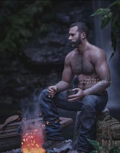 a shirtless man sitting next to a campfire with a cup in his hand