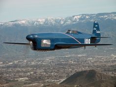 a blue airplane flying over a city with mountains in the background and snow capped peaks behind it