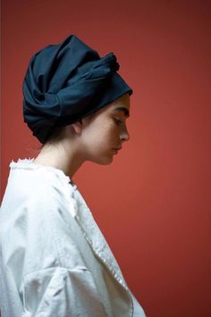 a woman wearing a black turban with her eyes closed, standing against a red wall